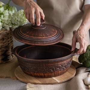 Puede incluir: Una olla de cerámica marrón con tapa, que presenta un diseño floral decorativo. La olla está sobre una tabla de cortar de madera.