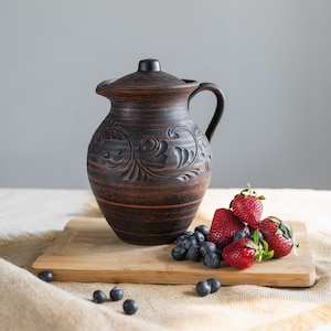 Könnte beinhalten: Ein brauner Keramik-Krug mit Deckel und aufwendigen Schnitzereien steht auf einem Holzbrett mit frischen Erdbeeren und Blaubeeren.