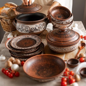 May include: A collection of handcrafted, rustic pottery. The set includes a large pot with a lid, bowls, plates, and a serving dish. The pottery is brown with white floral designs. The scene is set on a table with bread, tomatoes, and mushrooms.