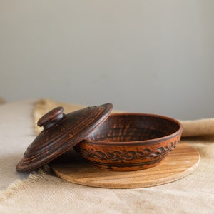 Op de afbeelding: Bruine keramische kom met deksel, op een houten plank. De kom heeft een decoratief patroon langs de rand. Het deksel is gedeeltelijk open. Ideaal voor het serveren van gerechten.