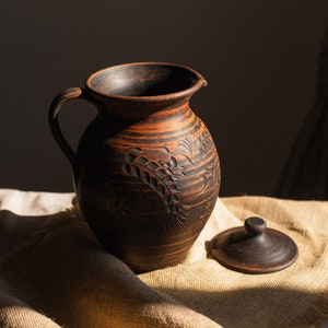 May include: A dark brown ceramic jug with a handle and a lid. The jug has a carved floral pattern and sits on a beige cloth. The jug is likely used for serving drinks. The jug is made of ceramic.