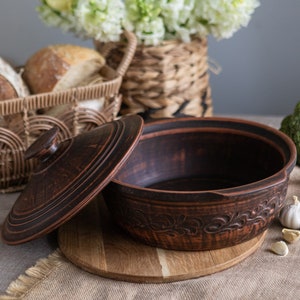 May include: A brown ceramic casserole dish with a lid, resting on a wooden cutting board. The dish has a decorative floral pattern around the rim. A basket of bread and flowers are in the background.