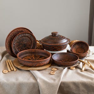 May include: A collection of rustic, terracotta-colored ceramic tableware. The set includes plates, bowls, and a lidded casserole dish, all with intricate embossed patterns. The items are arranged on a neutral-toned tablecloth, suggesting a warm, inviting aesthetic.