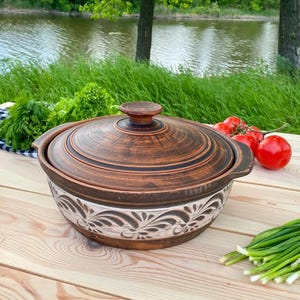Könnte beinhalten: Ein runder Kochtopf aus braunem Ton mit passendem Deckel, der oben ein dunkles, gewirbeltes Finish und an der Basis ein helleres, dekoratives Band mit geschnitzten Blumenmustern aufweist. Der Topf hat zwei kleine Seitengriffe.