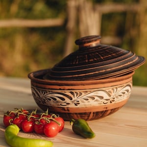 Op de afbeelding: Een bruine keramische ovenschaal met deksel, met een decoratief bloemmotief in wit en zwart. De schaal staat op een houten oppervlak met rode tomaten en een groene peper.