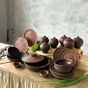 May include: A collection of rustic brown ceramic tableware, including plates, bowls, and lidded jars. The pottery has a textured finish and decorative patterns. The arrangement suggests a dining or serving set.