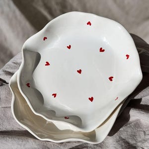 May include: Two white ceramic plates with wavy edges, each adorned with small red heart designs. The plates are stacked on top of each other, creating a layered effect. The plates are on a gray linen cloth.