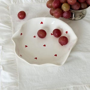 May include: A white ceramic dish with a wavy edge, decorated with small red hearts, holds several red grapes. A small silver bowl filled with grapes sits nearby. The dish and grapes rest on a white ruffled linen napkin.