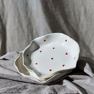May include: Two white ceramic plates with wavy edges, stacked on top of each other. Each plate is decorated with small, evenly spaced red hearts. The plates rest on a gray cloth, suggesting a kitchen or dining setting.