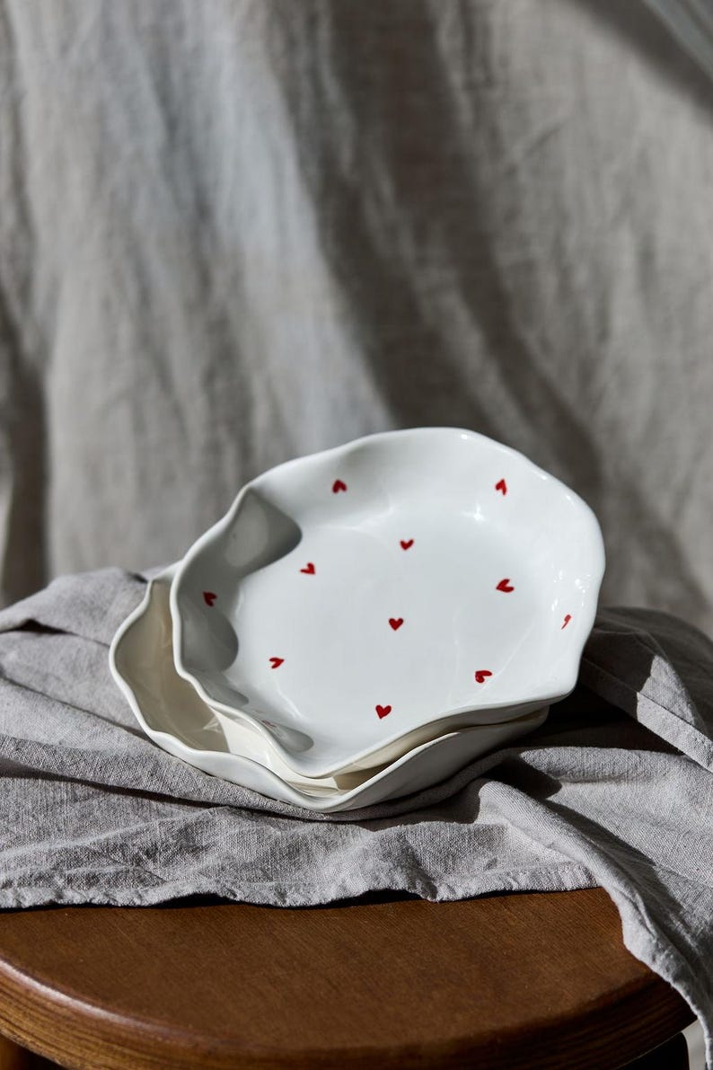 May include: Two white ceramic plates with wavy edges, stacked on top of each other. Each plate is decorated with small, evenly spaced red hearts. The plates rest on a gray cloth, suggesting a kitchen or dining setting.