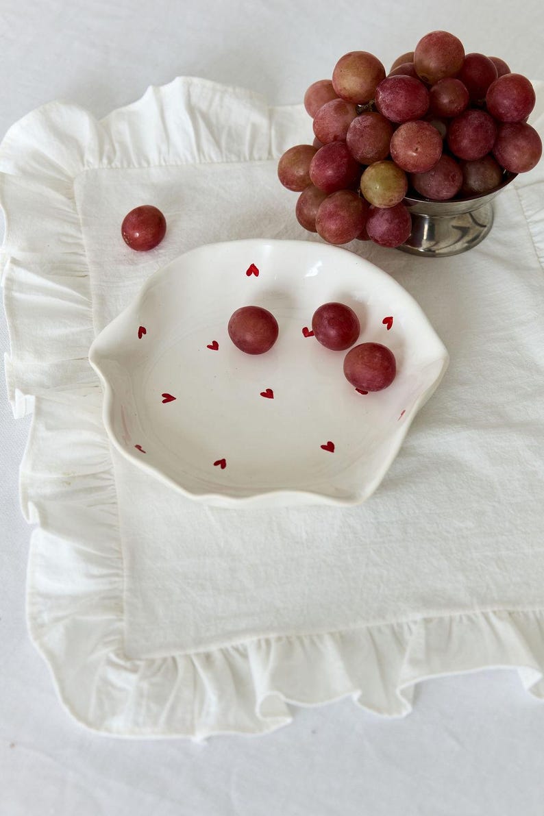 May include: A white ceramic dish with a wavy edge, decorated with small red hearts, holds several red grapes. A small silver bowl filled with grapes sits nearby. The dish and grapes rest on a white ruffled linen napkin.