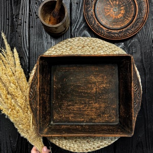 May include: A square dark brown clay baking dish with carved leaf patterns on the side handles, accompanied by a matching round plate, a wooden mortar and pestle, and a bundle of dried wheat stalks on a dark wooden surface.