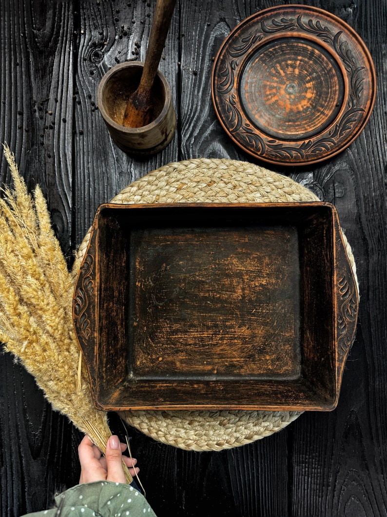 May include: A square dark brown clay baking dish with carved leaf patterns on the side handles, accompanied by a matching round plate, a wooden mortar and pestle, and a bundle of dried wheat stalks on a dark wooden surface.