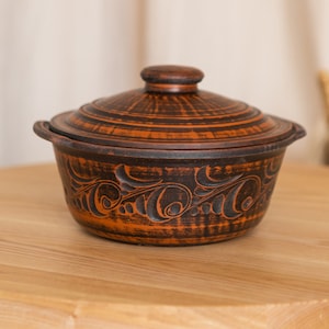 以下が含まれることがあります： 蓋付きの茶色の陶器製キャセロール皿。 皿は、オレンジ色のアクセントが付いた、暗い茶色の表面に彫られた装飾的な花の模様が特徴です。 明るい木製のテーブルの上に置かれています。