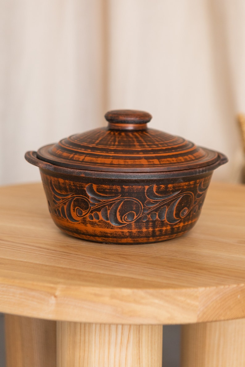 Może przedstawiać: Ręcznie wykonane, brązowe ceramiczne naczynie do zapiekania z pokrywką. Naczynie posiada skomplikowane, ciemne wzory kwiatowe wycięte na zewnątrz. Pokrywka ma mały, okrągły uchwyt. Naczynie stoi na drewnianym stole.