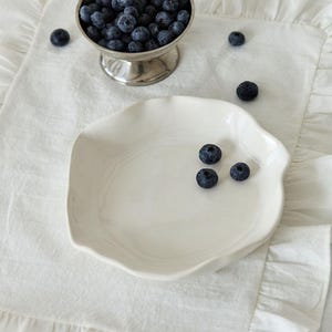 May include: A still life image featuring a silver footed bowl filled with fresh blueberries. A white, irregularly shaped plate holds a few blueberries, and the scene is set on a white, ruffled linen placemat.
