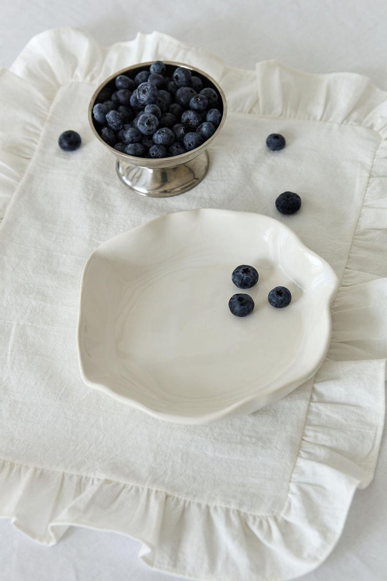 May include: A still life image featuring a silver footed bowl filled with fresh blueberries. A white, irregularly shaped plate holds a few blueberries, and the scene is set on a white, ruffled linen placemat.