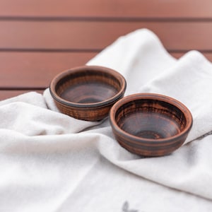 Puede incluir: Dos cuencos pequeños de cerámica marrón hechos a mano con interior oscuro y exterior texturizado. Los cuencos están colocados sobre un paño de lino blanco, creando una estética rústica y natural. Los cuencos probablemente se utilizan para servir salsas o porciones pequeñas.