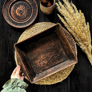 May include: A rectangular dark brown clay baking dish with carved leaf patterns on the handles, accompanied by a matching round plate, a small wooden mortar and pestle, and a bundle of dried beige pampas grass on a dark wooden surface.