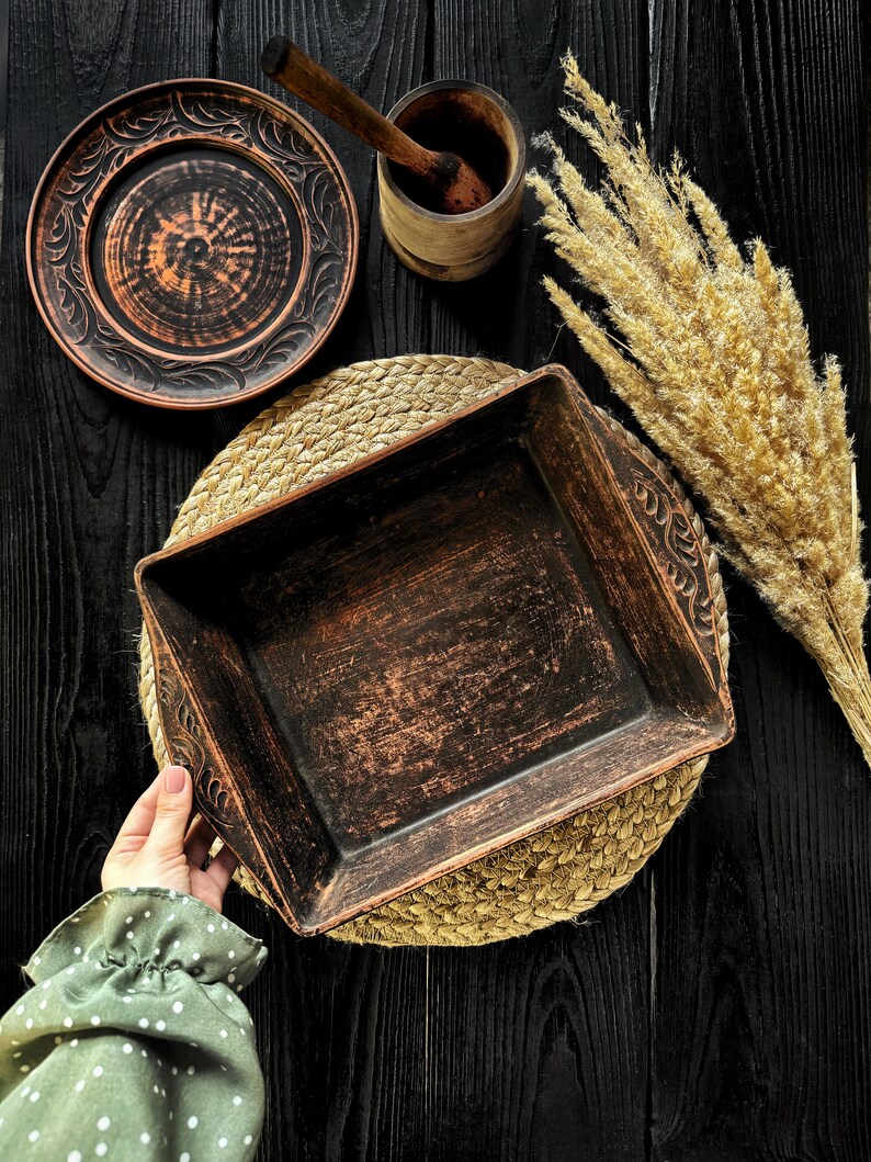 May include: A rectangular dark brown clay baking dish with carved leaf patterns on the handles, accompanied by a matching round plate, a small wooden mortar and pestle, and a bundle of dried beige pampas grass on a dark wooden surface.