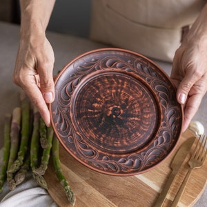 以下が含まれることがあります： 手作りの茶色のセラミックプレート。縁に精巧な浮き彫りの花柄模様があります。手で持たれており、背景にはアスパラガス、木製のまな板、金色のカトラリーがあります。直径は約25cmです。