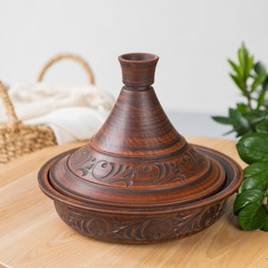 Puede incluir: Una tagine de cer&aacute;mica marr&oacute;n con intrincados dise&ntilde;os florales. La tagine est&aacute; sentada sobre una mesa de madera.