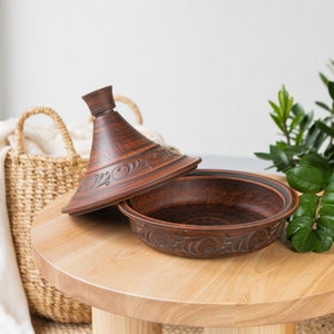 Puede incluir: Una tagine de cer&aacute;mica marr&oacute;n con intrincados tallados, abierta sobre una mesa de madera clara con una cesta tejida y una planta verde en el fondo.