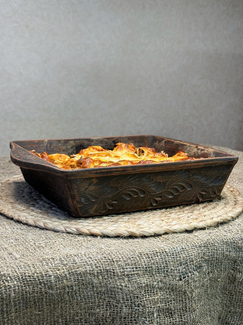 May include: A square, dark brown clay baking dish with an intricate, carved leaf pattern along the exterior sides. The dish is filled with a golden-brown, baked food item with a bubbly, melted cheese surface and visible browned edges.