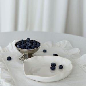 May include: A silver footed bowl filled with fresh blueberries sits on a white ruffled placemat next to a white ceramic dish. Several blueberries are scattered around the bowl and dish. The background is a white curtain.