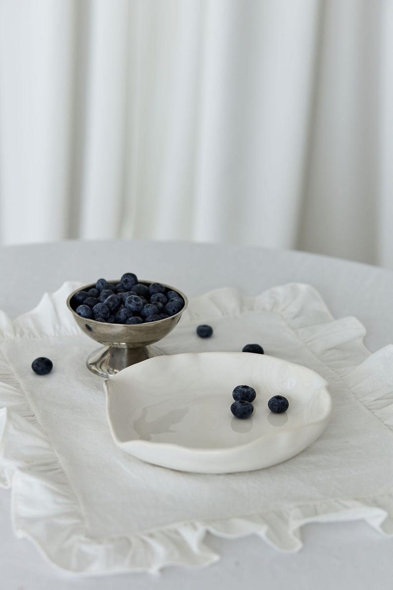 May include: A silver footed bowl filled with fresh blueberries sits on a white ruffled placemat next to a white ceramic dish. Several blueberries are scattered around the bowl and dish. The background is a white curtain.