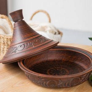 Puede incluir: Una tagine de cer&aacute;mica marr&oacute;n con tapa, que presenta intrincados dise&ntilde;os florales. La tagine est&aacute; sentada sobre una mesa de madera.