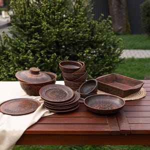 Könnte beinhalten: Eine Sammlung brauner Keramikgeschirr, darunter Teller, Schalen und eine Auflaufform mit Deckel, auf einem Holztisch angeordnet. Die Stücke weisen aufwendige Schnitzereien auf und sind vor Grün angeordnet.