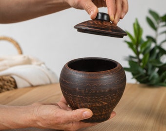May include: A handmade, dark brown ceramic pot with a lid. The pot has a rounded shape and is decorated with etched patterns. The lid is being lifted, revealing the interior. The pot is held by hands, suggesting its size and use.