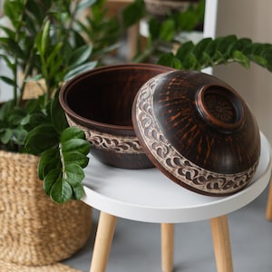 May include: A white ceramic mug with a black handle and the text "But First, Coffee" in a bold, black font. The mug is set against a blurred background, suggesting a cosy setting.