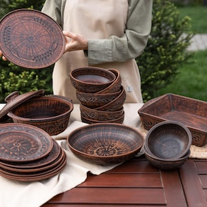 May include: A collection of dark brown terracotta kitchenware including a stack of plates, a lidded pot, several bowls, and a rectangular baking dish. Each piece features intricate embossed patterns and a rustic, unglazed clay texture throughout.
