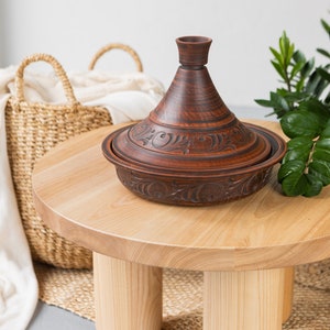 Puede incluir: Una tagine de cer&aacute;mica marr&oacute;n con intrincados dise&ntilde;os florales se encuentra sobre una mesa de madera clara con tres patas. La tagine es una olla de cocci&oacute;n tradicional marroqu&iacute; con una tapa c&oacute;nica.