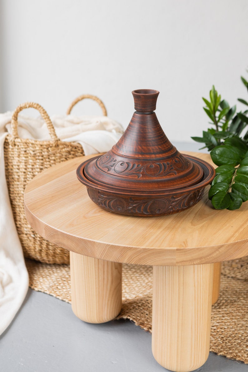 Puede incluir: Una tagine de cer&aacute;mica marr&oacute;n con intrincados dise&ntilde;os florales se encuentra sobre una mesa de madera clara con tres patas. La tagine es una olla de cocci&oacute;n tradicional marroqu&iacute; con una tapa c&oacute;nica.