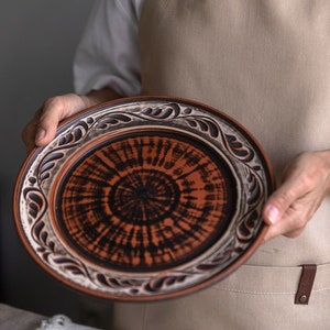 May include: A brown ceramic plate with a white rim and black floral design. The plate has a circular, swirling pattern in the center.