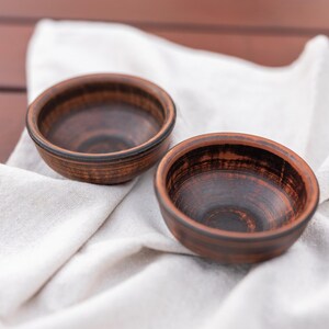 May include: Two small, handmade ceramic bowls with a dark brown exterior and interior. The bowls are set on a white linen cloth, creating a rustic and natural aesthetic. The bowls are ideal for serving dips, snacks, or small portions.
