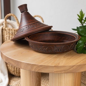 Puede incluir: Una tagine de barro marr&oacute;n con tapa, sentada sobre una mesa de madera clara con tres patas. La tagine tiene un dise&ntilde;o floral decorativo.