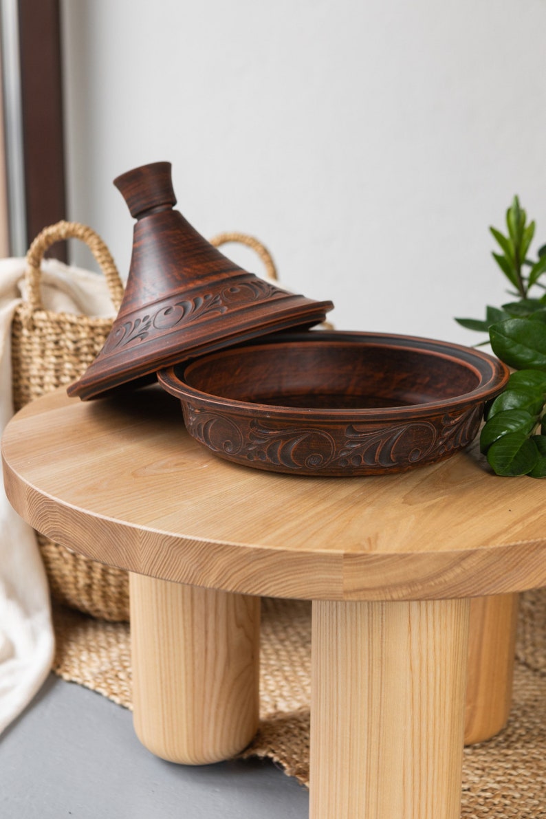 Puede incluir: Una tagine de barro marr&oacute;n con tapa, sentada sobre una mesa de madera clara con tres patas. La tagine tiene un dise&ntilde;o floral decorativo.