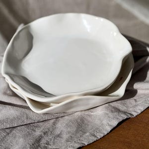 May include: Two white ceramic plates with irregular, wavy edges are stacked on a gray linen cloth. The plates have a glossy finish and a simple, elegant design. The plates are suitable for serving food.