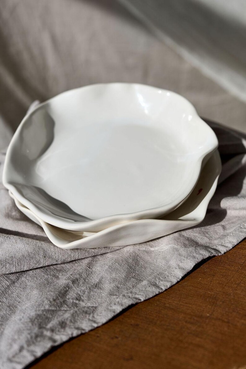 May include: Two white ceramic plates with irregular, wavy edges are stacked on a gray linen cloth. The plates have a glossy finish and a simple, elegant design. The plates are suitable for serving food.