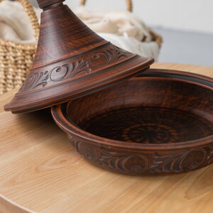 Puede incluir: Una tagine de barro marr&oacute;n con tapa, que presenta intrincados dise&ntilde;os florales. La tagine est&aacute; sentada sobre una mesa de madera.