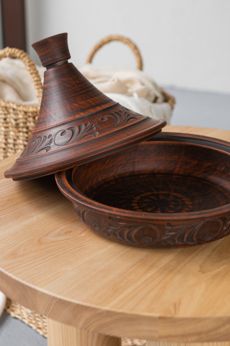 Puede incluir: Una tagine de barro marr&oacute;n con tapa, que presenta intrincados dise&ntilde;os florales. La tagine est&aacute; sentada sobre una mesa de madera.