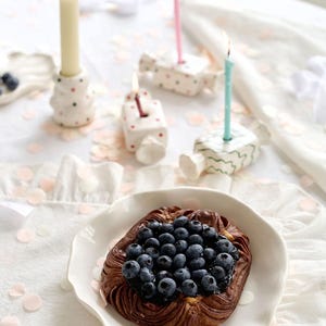May include: A baked pastry topped with fresh blueberries sits on a white plate. Lit candles in decorative holders and confetti are arranged on a white tablecloth, creating a festive scene. The pastry is the focal point.