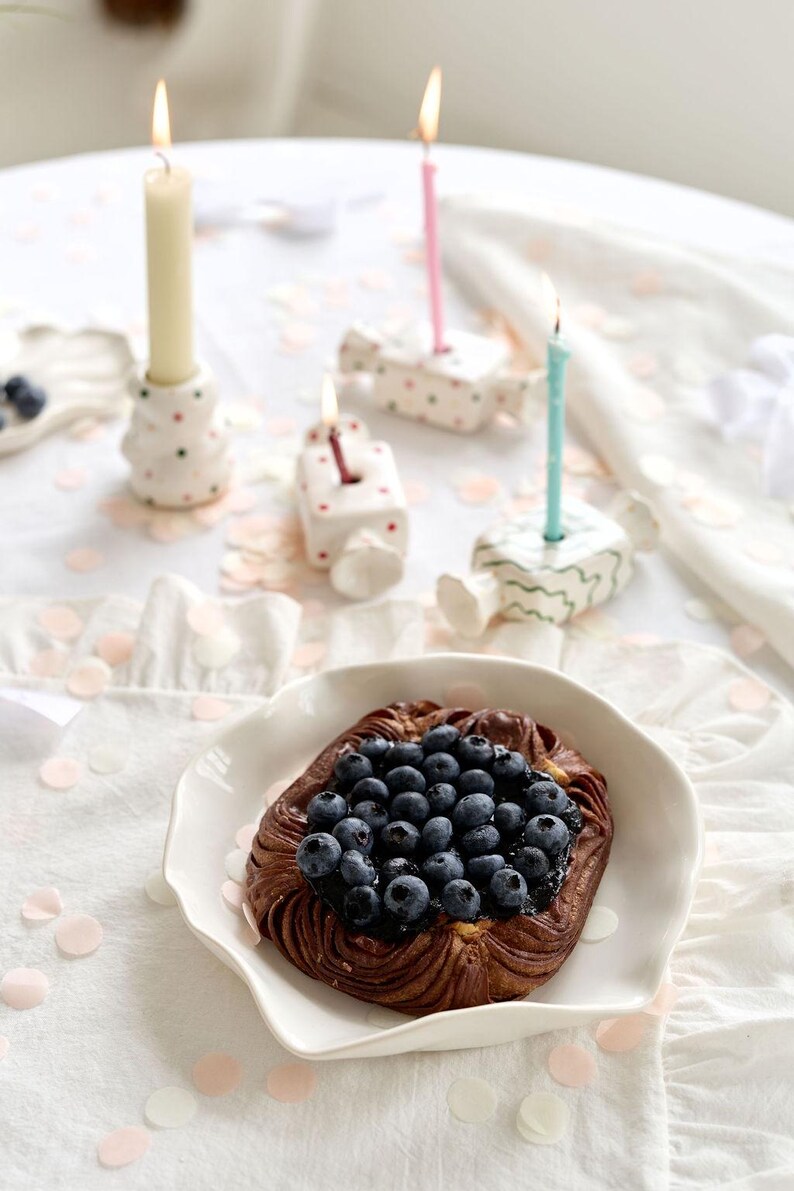 May include: A baked pastry topped with fresh blueberries sits on a white plate. Lit candles in decorative holders and confetti are arranged on a white tablecloth, creating a festive scene. The pastry is the focal point.
