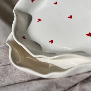 May include: Two white ceramic plates with a wavy edge and red heart accents. The plates are stacked on top of each other, with the top plate in focus. The plates are on a gray linen surface.