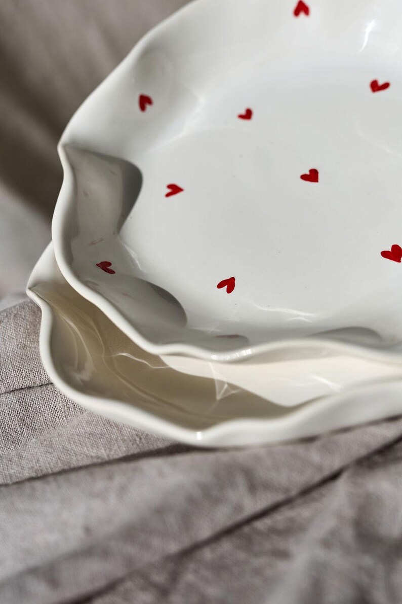 May include: Two white ceramic plates with a wavy edge and red heart accents. The plates are stacked on top of each other, with the top plate in focus. The plates are on a gray linen surface.