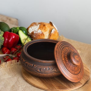 以下が含まれることがあります： 茶色のセラミックボウルと蓋。縁には渦巻き模様が施されています。蓋は部分的に開いており、暗い内部が見えます。ボウルは木製のまな板の上に置かれ、背景には新鮮な野菜とパンがあります。